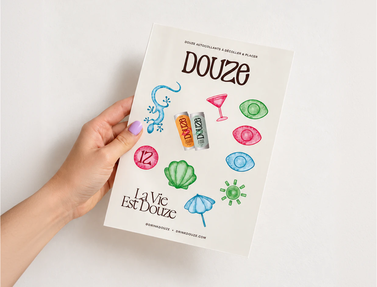 A person holding a white sticker sheet featuring the title 'DOUZE' and colorful illustrations of drinks, a lizard, a seashell, and eyes