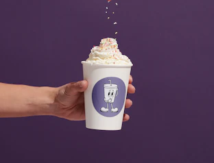 A hand holding a white paper cup topped with whipped cream and colorful sprinkles against a purple background