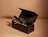 A dark wine bottle lying in an open black hinged presentation box filled with paper shred on a beige background