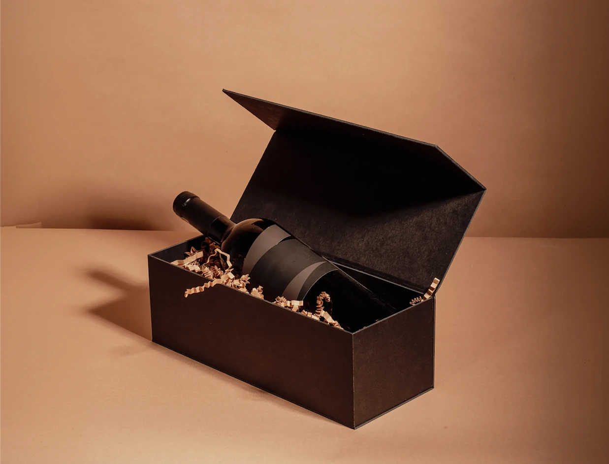 A dark wine bottle lying in an open black hinged presentation box filled with paper shred on a beige background