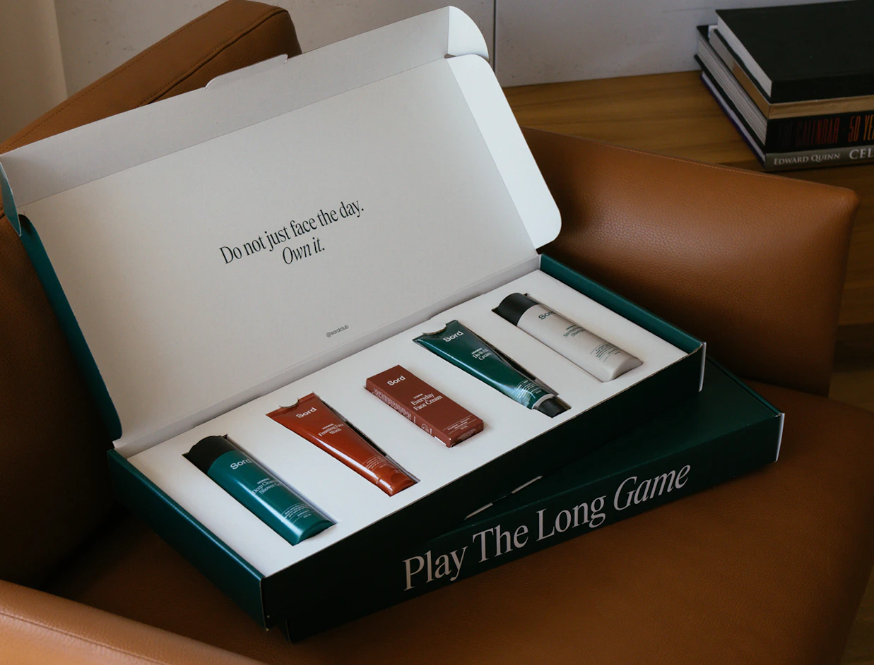 An open dark green gift box on a leather chair displaying five Sord men's skincare products in custom slots