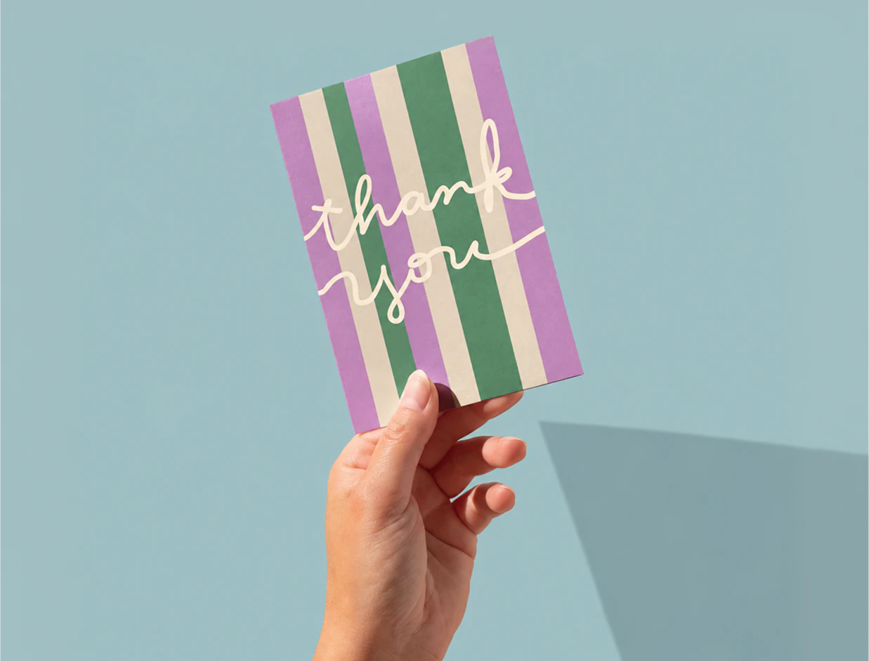 A hand holding a striped purple and green card with handwritten text 'thank you' against a blue background