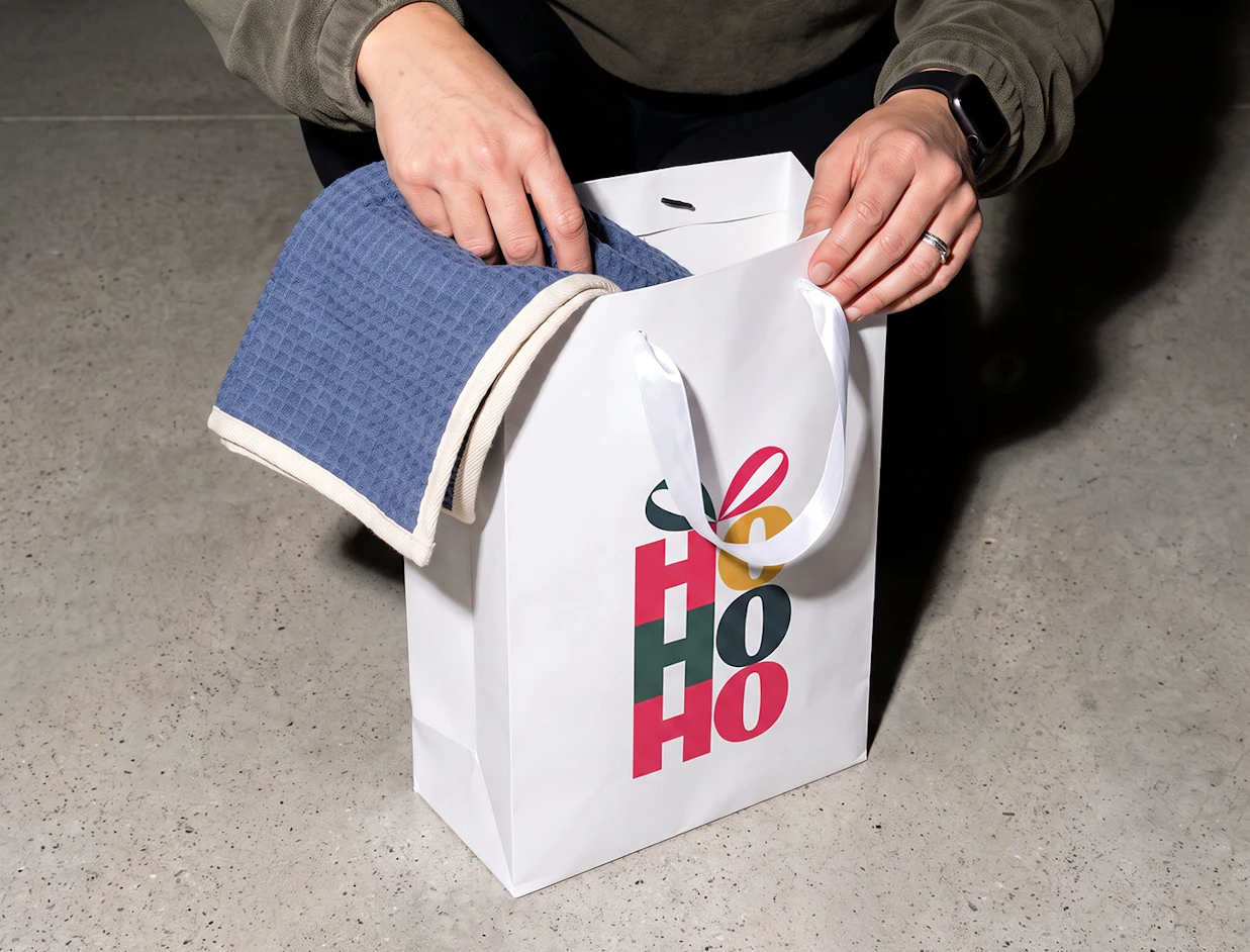 A person placing a blue waffle towel into a white gift bag printed with festive 'HO HO HO' typography