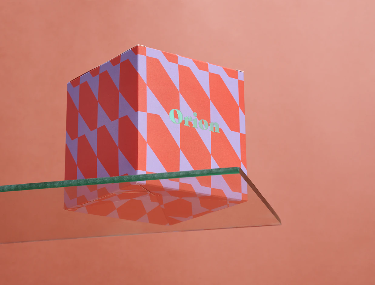 A red and purple geometric patterned box labeled 'Orion' sitting on a clear glass shelf with reflection