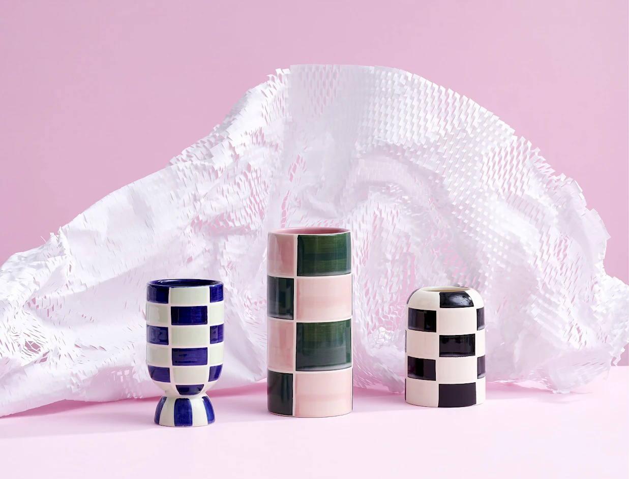 Three checkered ceramic vases standing in front of expanded white honeycomb packaging paper on a pink background