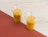 Two clear plastic cups with orange juice and glass straws, labeled 'Jaglo' and 'JOHN COFFE', placed on a wooden and red surface