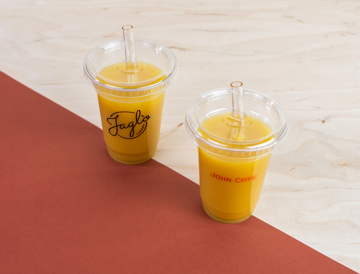 Two clear plastic cups with orange juice and glass straws, labeled 'Jaglo' and 'JOHN COFFE', placed on a wooden and red surface