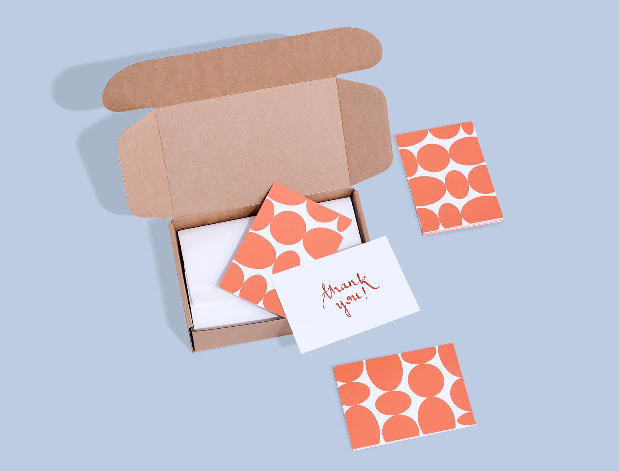 An open cardboard shipping box with patterned orange insert cards and a white 'thank you!' note