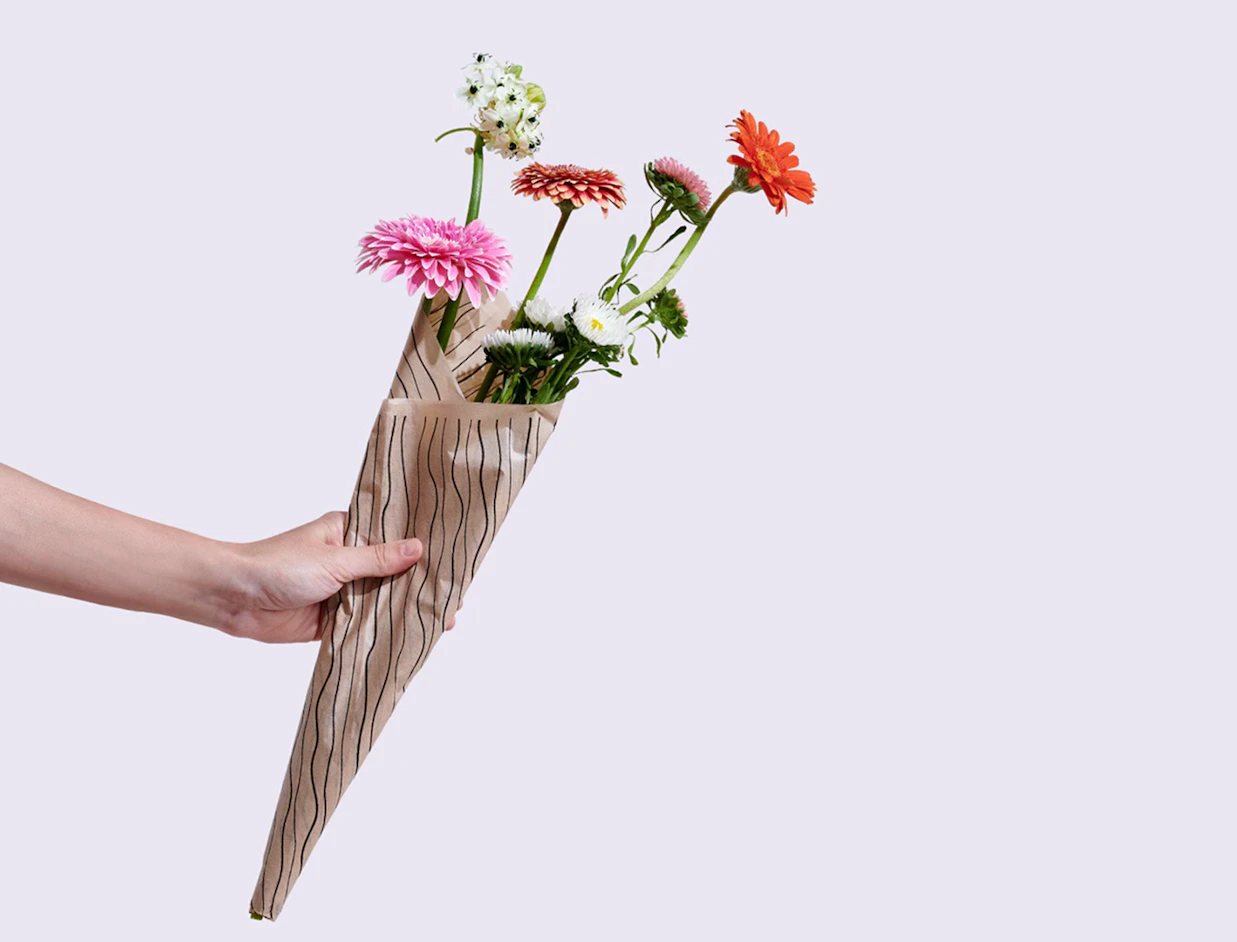 A hand holding a flower bouquet wrapped in brown paper with black wavy lines on a plain light background
