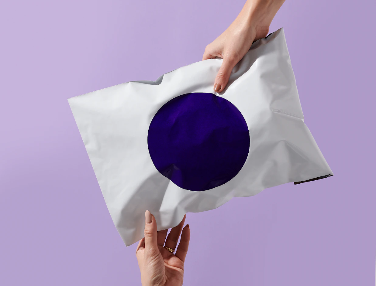 Two hands holding a white poly mailer with a large dark purple circle printed in the center against a light purple background