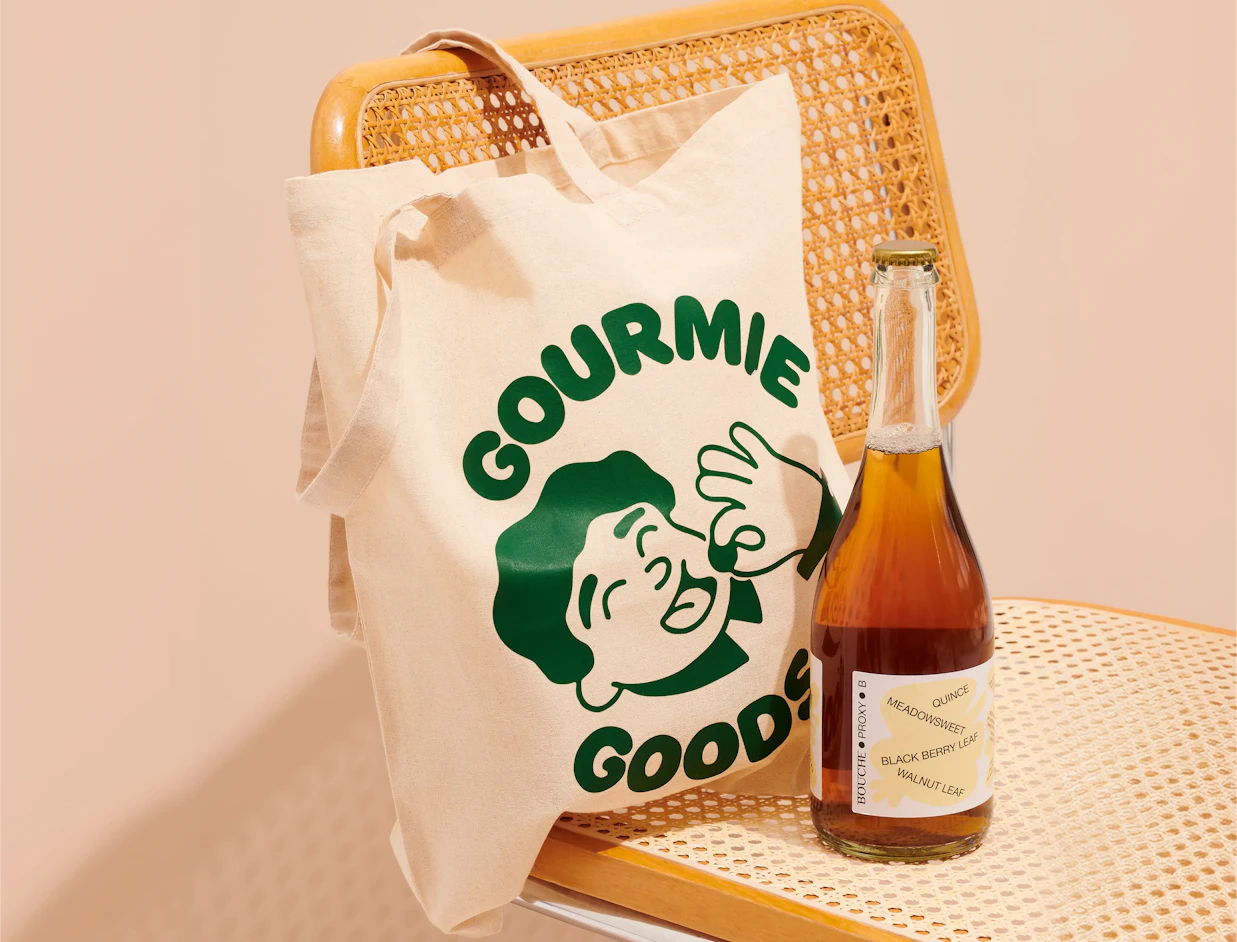 Natural cotton tote bag with a green illustrated logo print, hanging on a rattan chair
