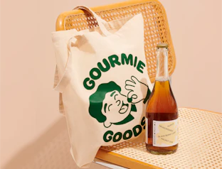 Natural cotton tote bag with a green illustrated logo print, hanging on a rattan chair