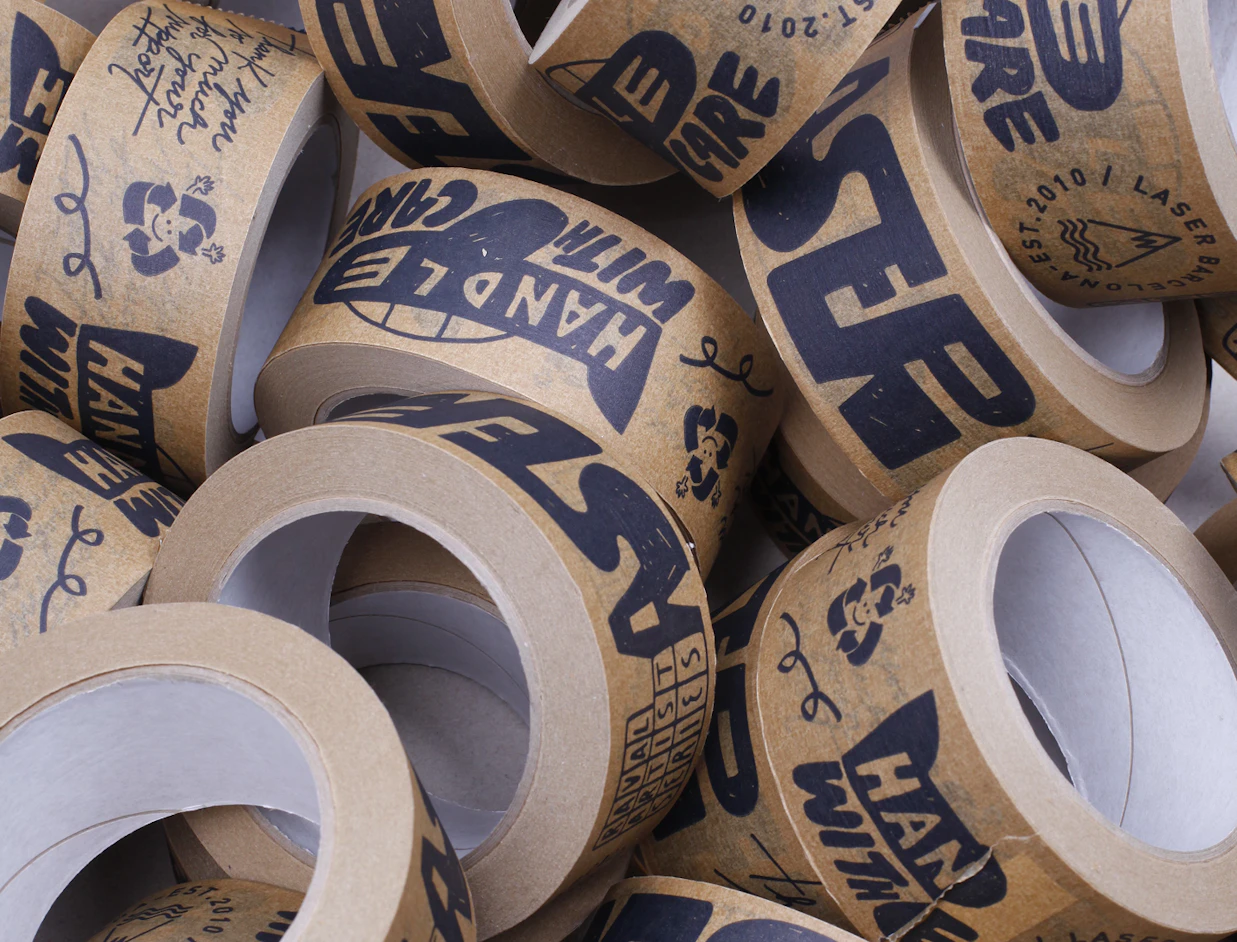 Close-up pile of brown paper tape rolls printed with 'HANDLE WITH CARE' and recycling icons in dark ink