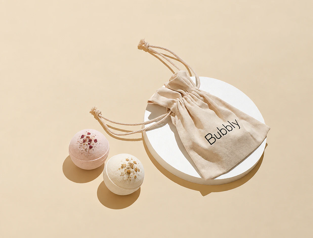 A beige drawstring cotton pouch labeled 'Bubbly' on a white circular display stand next to two round bath bombs
