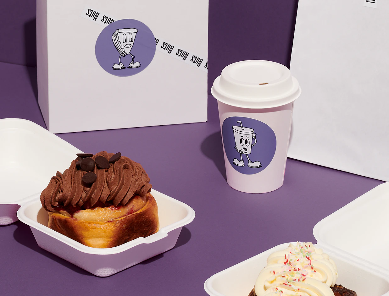 Pastry boxes and a paper cup with cartoon stickers next to chocolate and cream buns on a purple surface