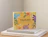A yellow mailer box decorated with colorful botanical illustrations and 'YOUR DAY just got a lot better!' text resting on a stack of books