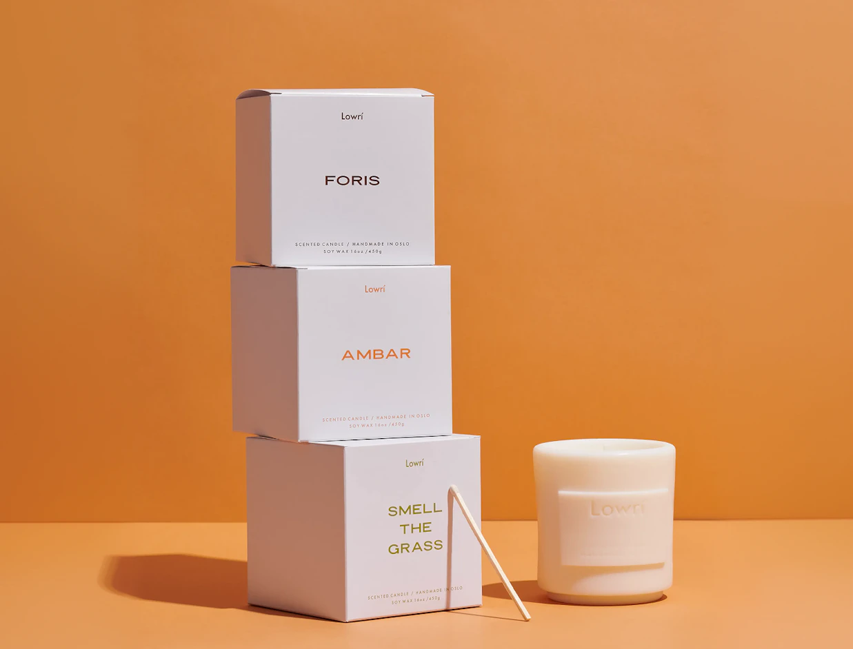 Three stacked white Lowrí candle boxes next to an unlit white candle and a match stick on an orange background
