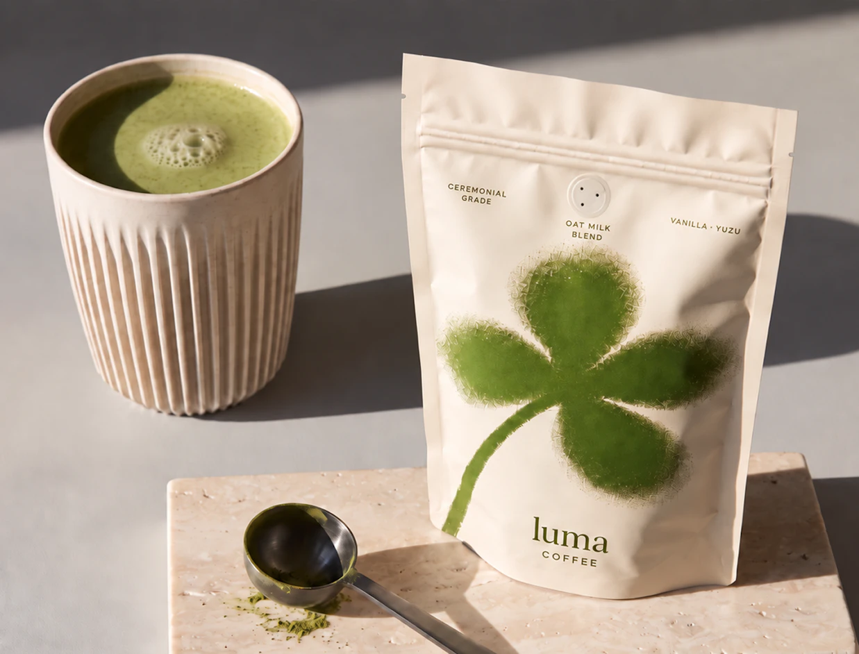A cream-colored ziplock pouch printed with a green four-leaf clover logo branded 'luma COFFEE' next to a cup of matcha latte and a metal scoop