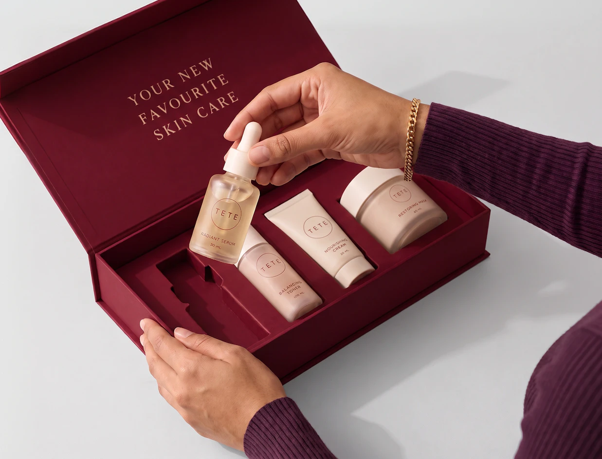 Hands opening a deep red rigid box with custom molded inlay holding TETE skincare bottles and jars, with 'YOUR NEW FAVOURITE SKIN CARE' printed inside the lid