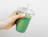 Hand holding a clear plastic cup with green smoothie and a glass straw, branded with the green 'Randy's' logo, tilted against a white background