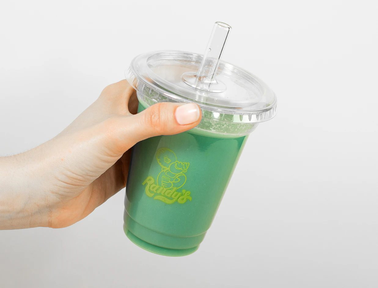 Hand holding a clear plastic cup with green smoothie and a glass straw, branded with the green 'Randy's' logo, tilted against a white background