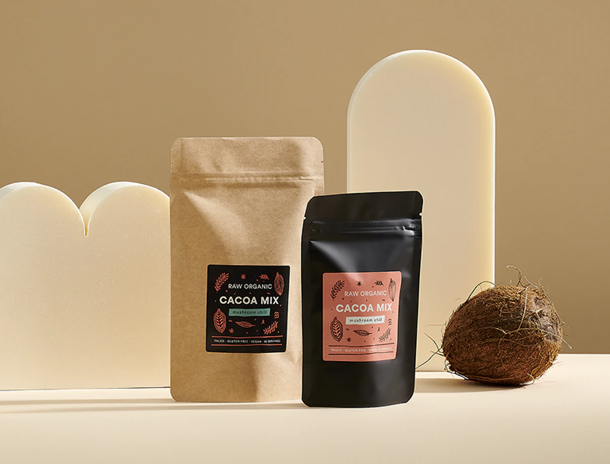 Two 'RAW ORGANIC CACOA MIX' pouches in kraft brown and matte black set against cream geometric props with a whole coconut