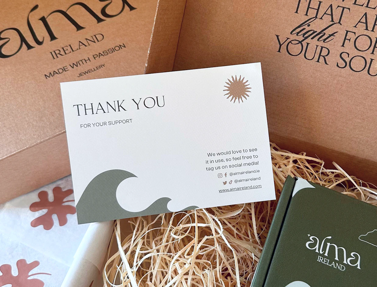 A white thank you card with a wave design placed inside a box filled with wood wool, branded with 'alma IRELAND'