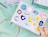 Hand holding a purple sticker sheet with various colorful space, dinosaur, and retro-themed stickers
