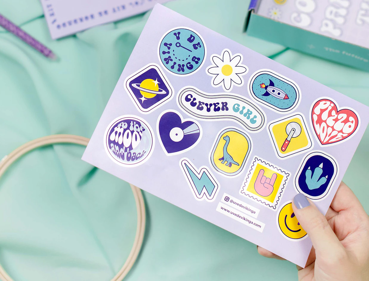 Hand holding a purple sticker sheet with various colorful space, dinosaur, and retro-themed stickers