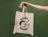 Hand holding a natural cotton tote bag with a green illustrated print against a green wall