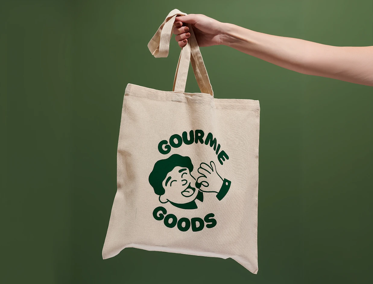 Hand holding a natural cotton tote bag with a green illustrated print against a green wall