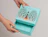 Hands opening a light blue box decorated with an ornate metallic peacock illustration labeled 'PAÓ GIN'