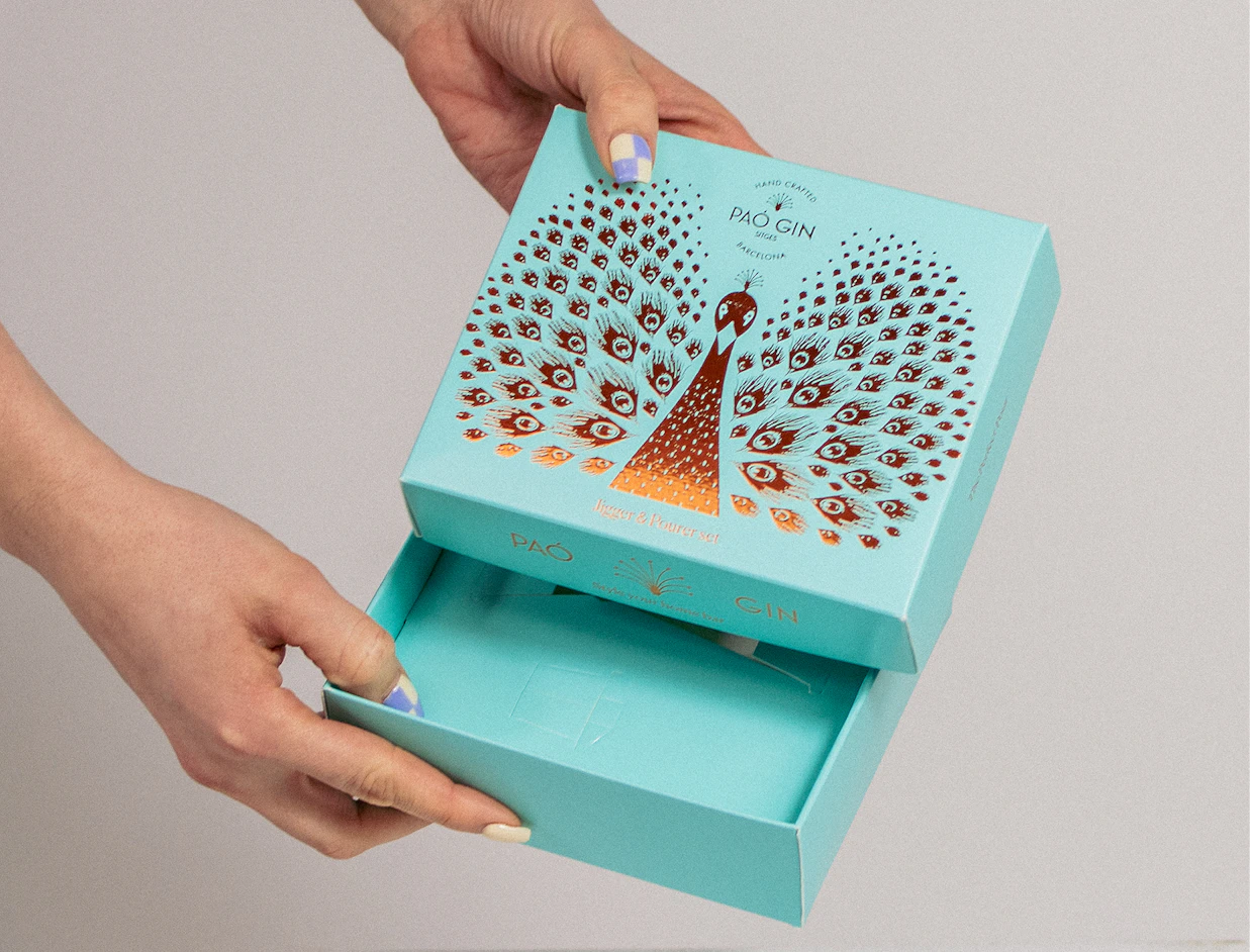 Hands opening a light blue box decorated with an ornate metallic peacock illustration labeled 'PAÓ GIN'