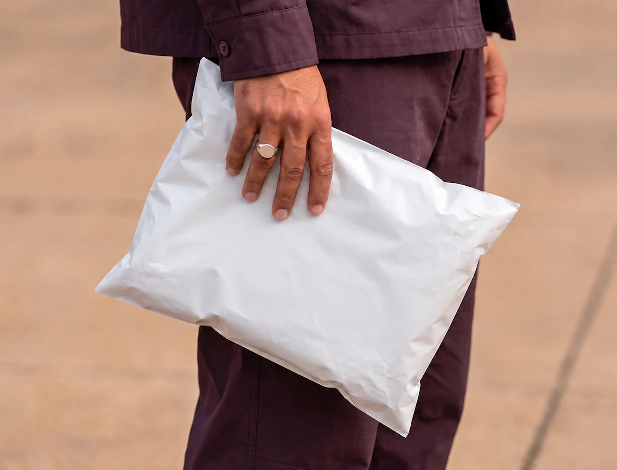 A person in a dark outfit holding a plain white poly mailer bag against a neutral background