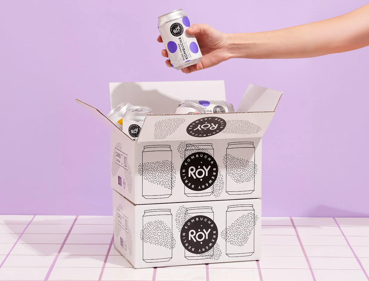 Hand taking a can from an open white shipping box with a black print on a lilac background