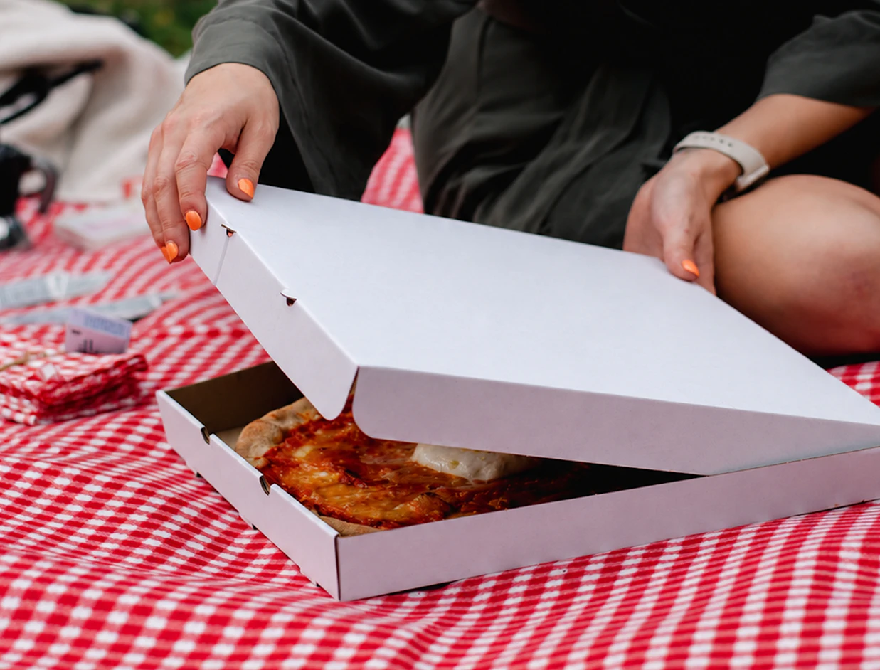 Hands opening a plain white cardboard pizza box revealing a fresh pizza on a red and white checkered picnic blanket