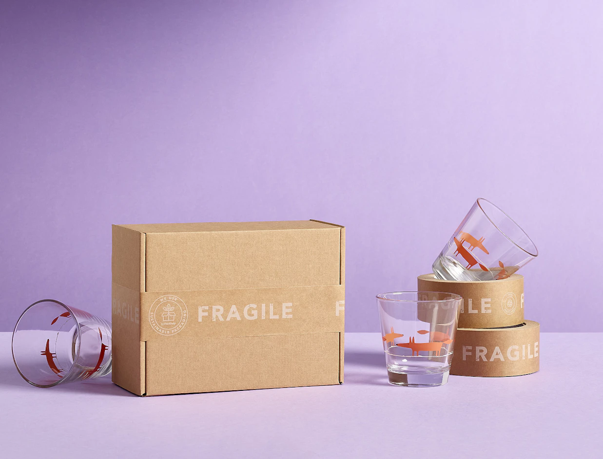 A cardboard box sealed with 'FRAGILE' paper tape surrounded by glass cups printed with red fox illustrations on a purple background