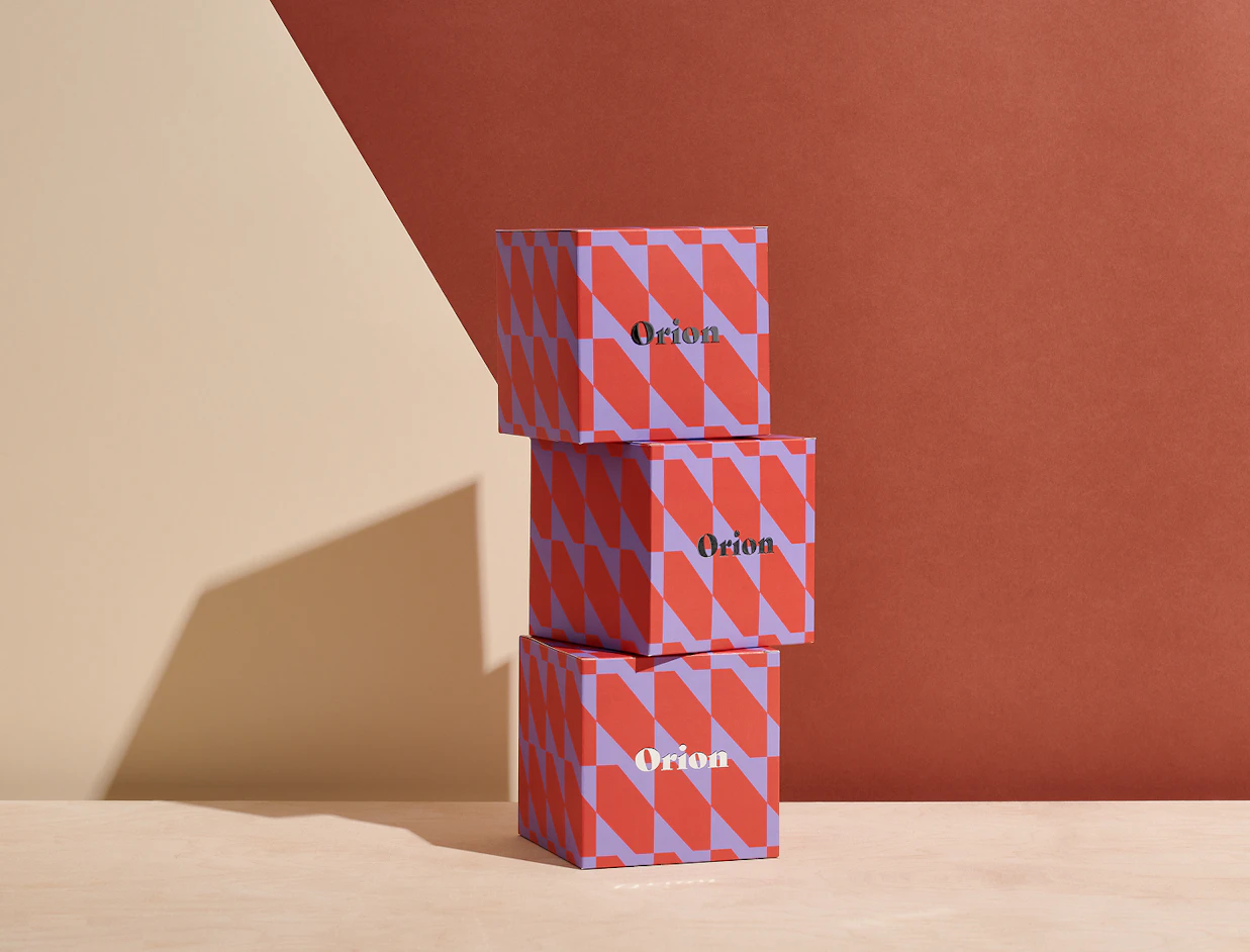 Three red and purple geometric patterned boxes labeled 'Orion' stacked vertically on a wooden table