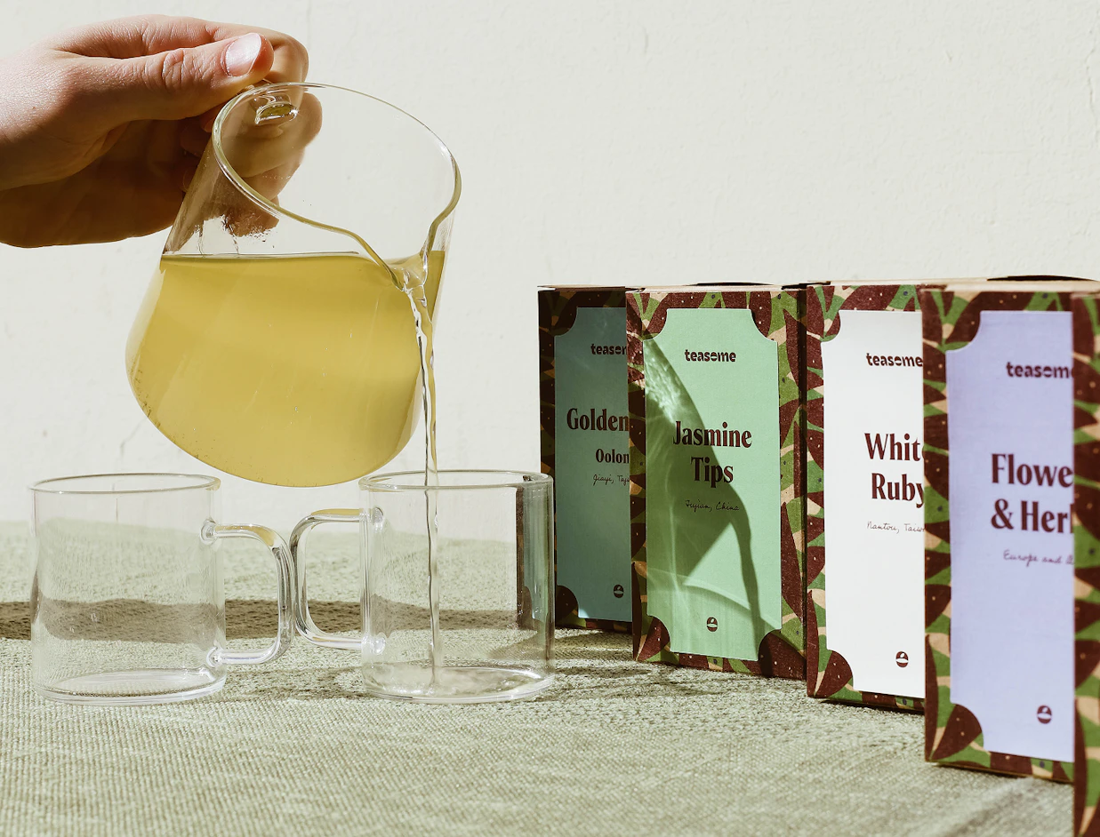 A hand pouring tea from a glass pitcher into a glass cup next to four colorful 'teaseme' tea packaging boxes on a table