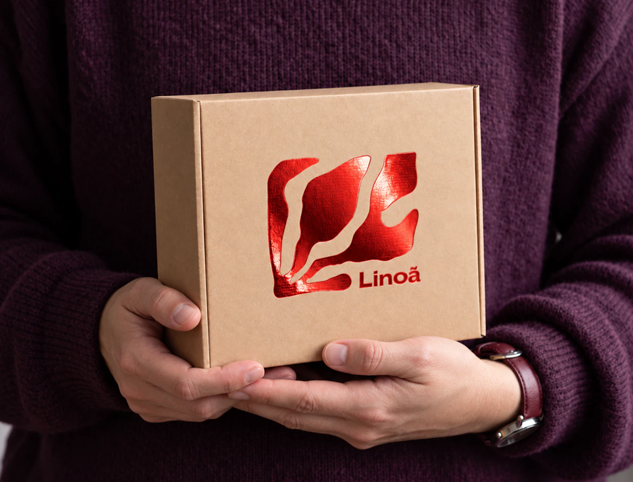Hands holding a kraft cardboard box embellished with a shiny red metallic foil 'Linoã' logo, set against a dark purple sweater
