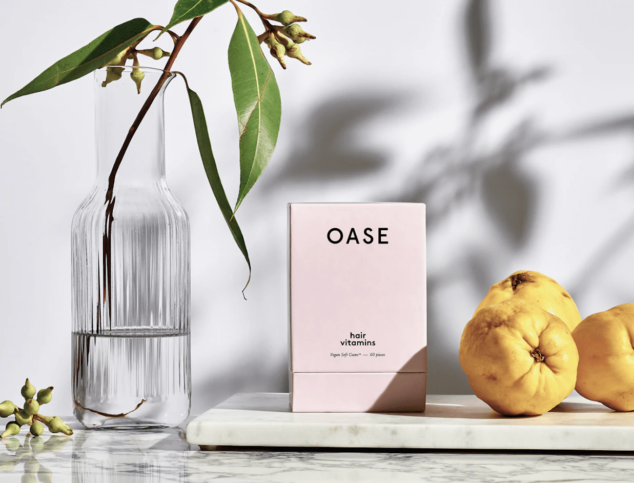A light pink OASE hair vitamins box on a marble slab next to yellow quinces and a glass vase with leaves