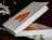 A partially open white pizza box with an orange tomato graphic on top, displaying a pizza inside and social media handles on the side