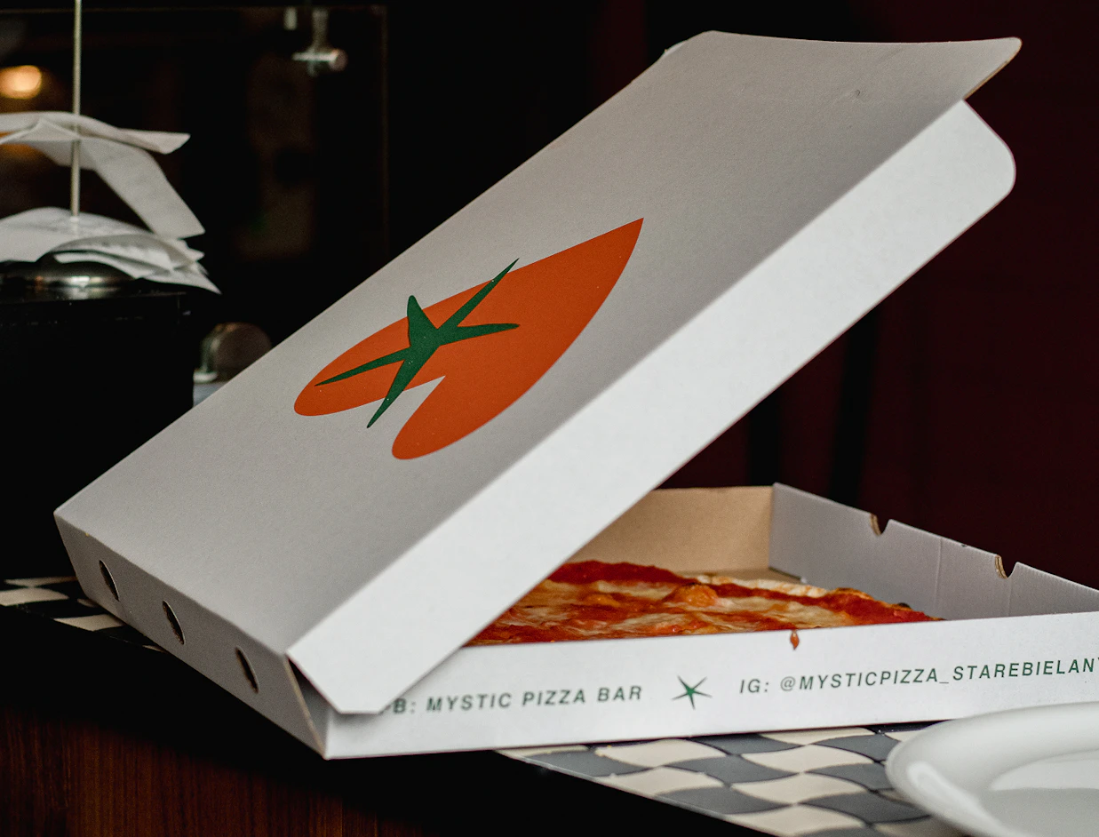 A partially open white pizza box with an orange tomato graphic on top, displaying a pizza inside and social media handles on the side