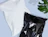 Top-down view of a plain white poly mailer and a glossy black poly mailer overlapping on a light blue surface next to a monstera leaf