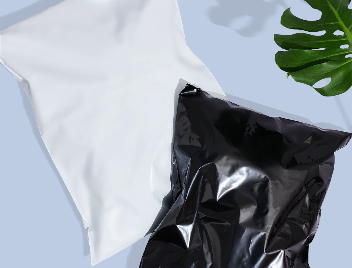 Top-down view of a plain white poly mailer and a glossy black poly mailer overlapping on a light blue surface next to a monstera leaf