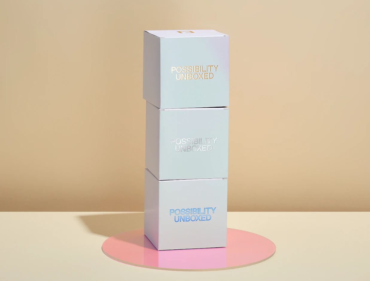 Three light blue cube boxes stacked vertically on a pink acrylic circle, each printed with 'POSSIBILITY UNBOXED' in metallic foil finish