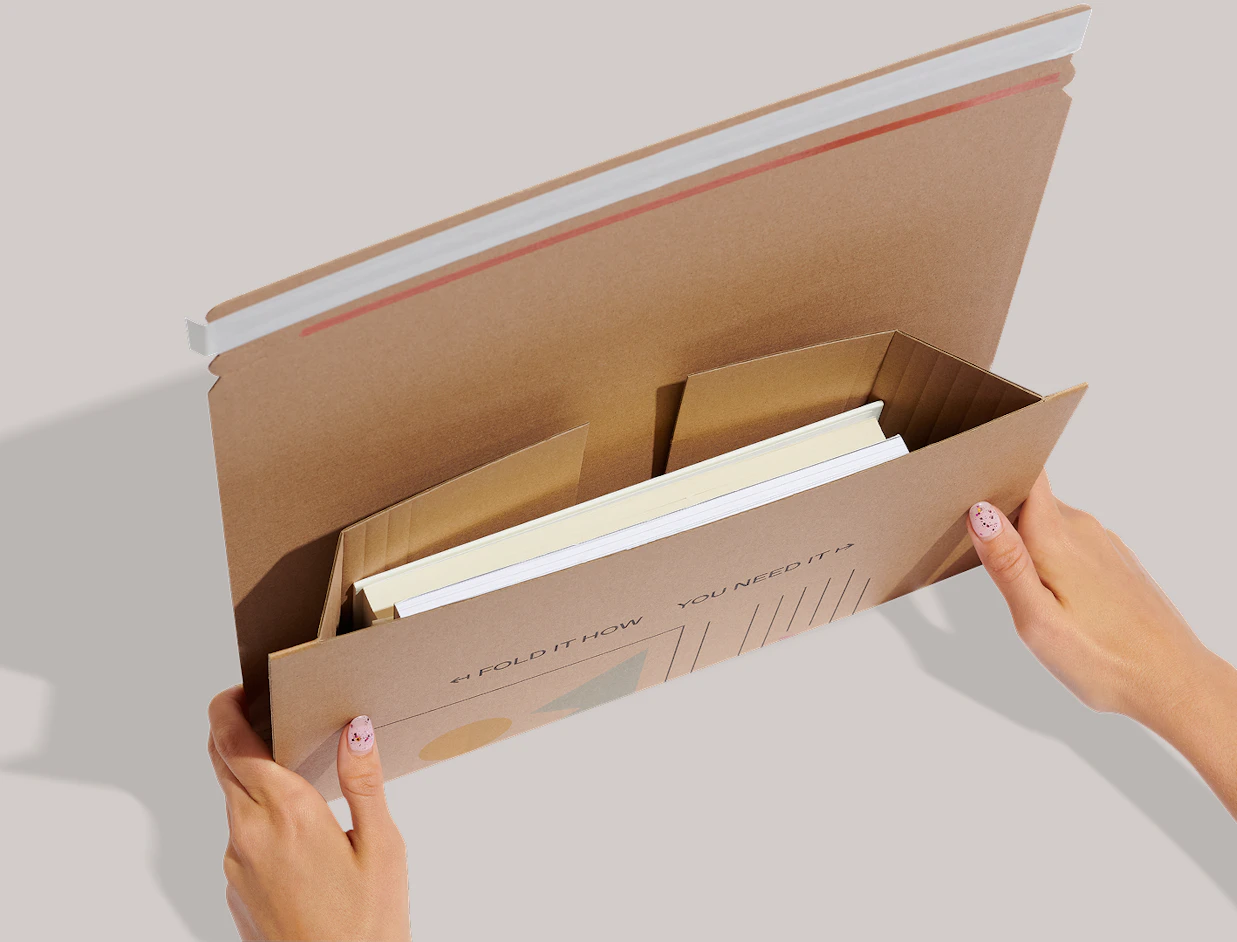 Hands holding open a cardboard book mailer showing books tucked inside internal flaps, printed with 'FOLD IT HOW YOU NEED IT'