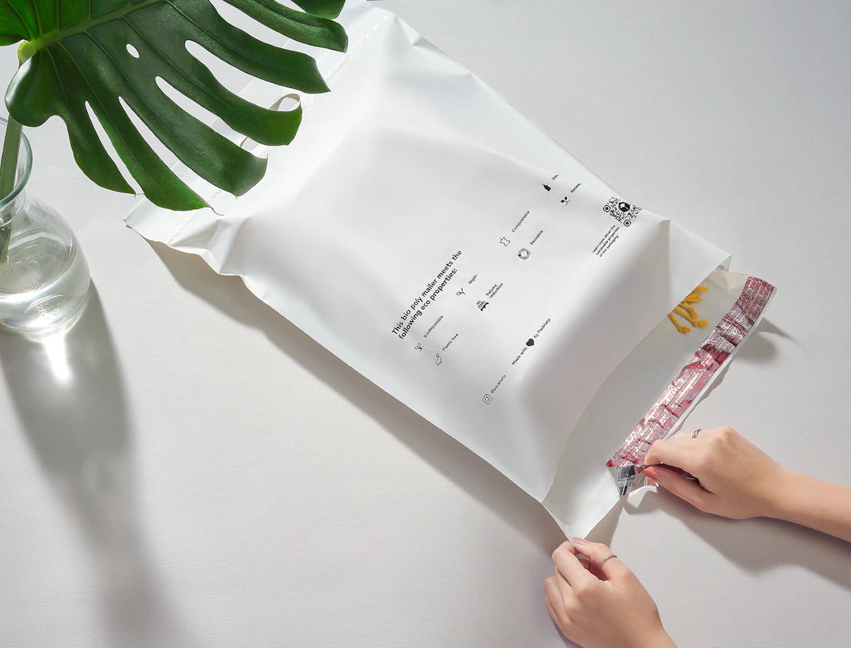 Hands peeling the red protective strip off a white bio poly mailer bag printed with sustainability icons, placed next to a monstera leaf