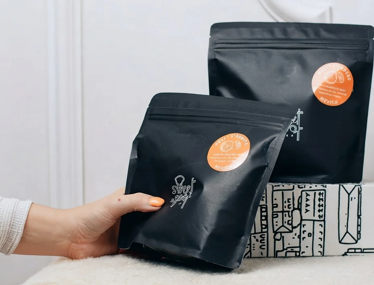 A hand holding a matte black ziplock coffee pouch with an orange round label, displayed in front of another black pouch and a patterned box