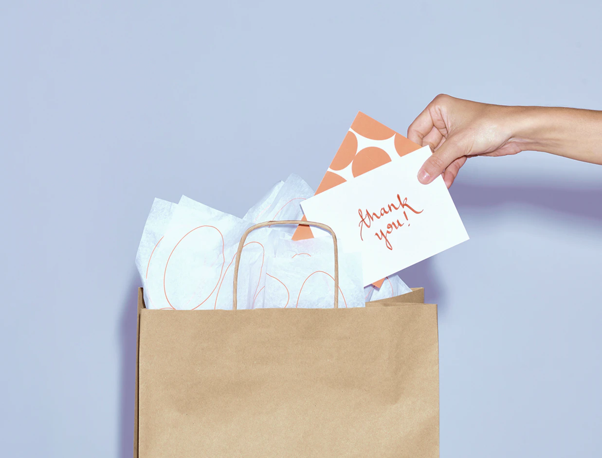 A hand pulling a white 'thank you!' card out of a brown paper shopping bag filled with tissue paper